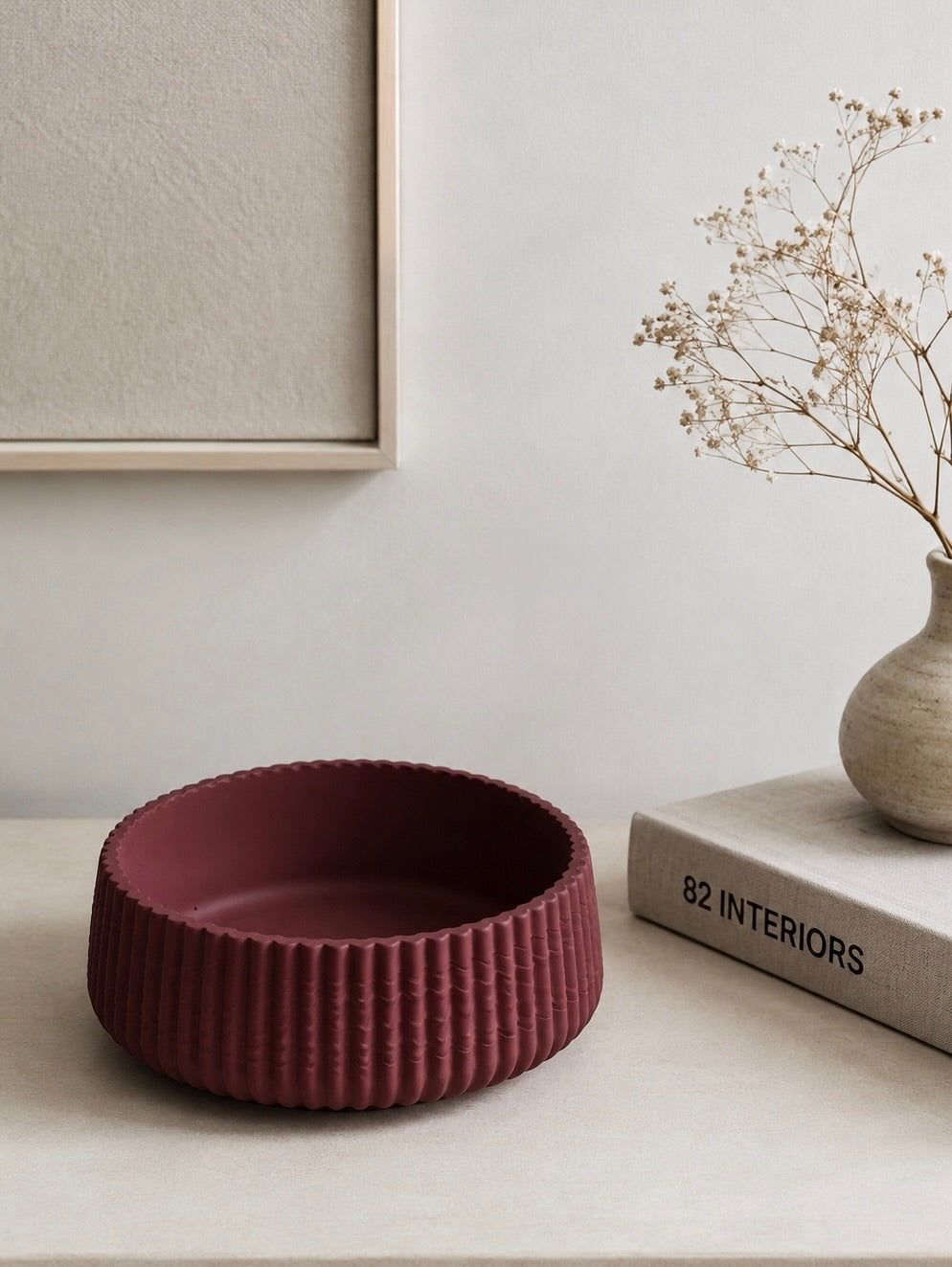 A textured, ribbed jesmonite bowl in a deep burgundy red sits on a light beige tabletop. The background features a neutral wall with a framed print, a small vase with dried flowers, and a book titled '82 INTERIORS'.