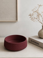 A textured, ribbed jesmonite bowl in a deep burgundy red sits on a light beige tabletop. The background features a neutral wall with a framed print, a small vase with dried flowers, and a book titled '82 INTERIORS'.