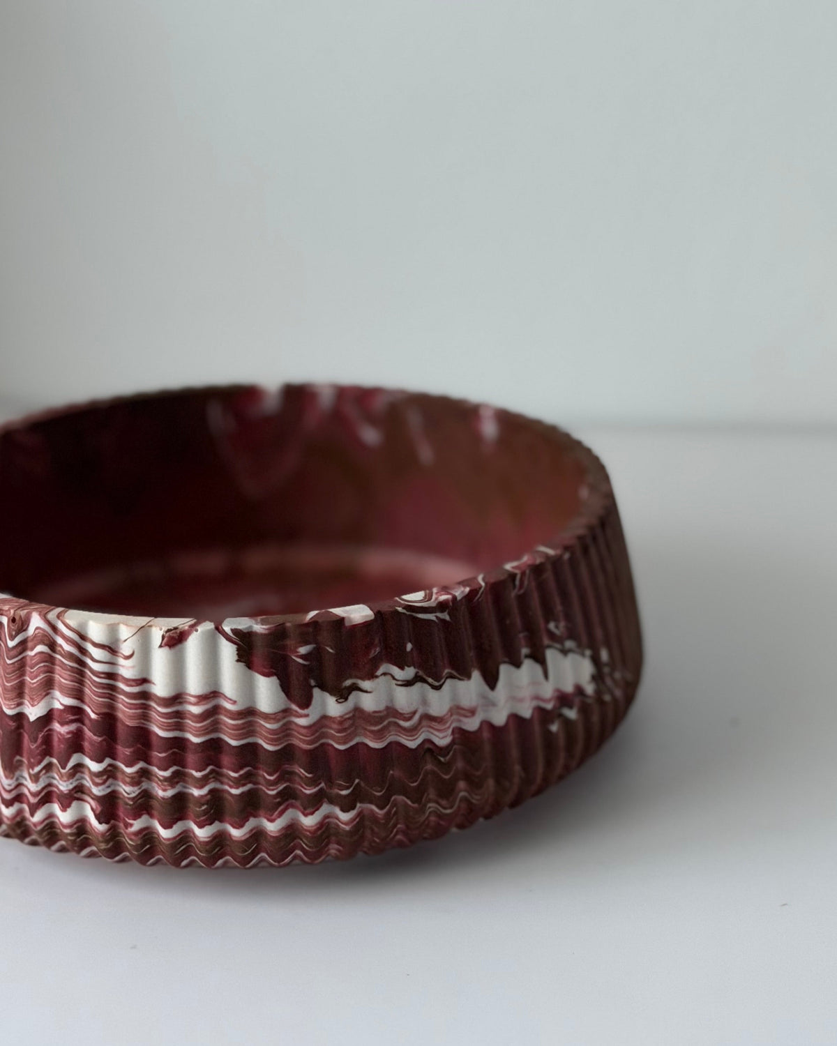 A handmade ceramic bowl with a deep maroon-brown glaze and a textured, ridged exterior featuring a wavy white and dark pattern, photographed on a plain light gray surface.