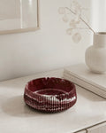 A ribbed, deep burgundy marbled jesmonite handmade bowl with a stone finish, styled on a light beige console table next to a white ceramic vase with dried leaves.