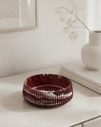 A ribbed, deep burgundy marbled jesmonite handmade bowl with a stone finish, styled on a light beige console table next to a white ceramic vase with dried leaves.