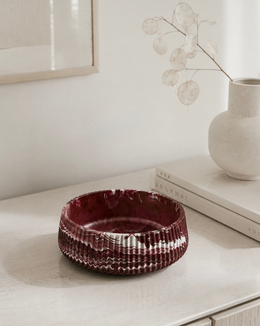 A ribbed, deep burgundy marbled jesmonite handmade bowl with a stone finish, styled on a light beige console table next to a white ceramic vase with dried leaves.